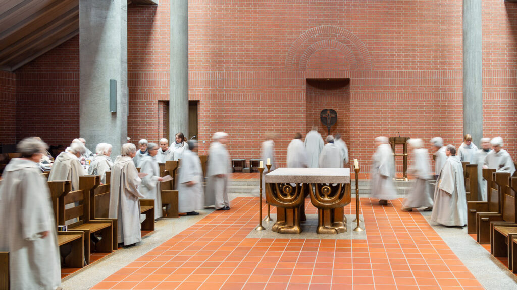 Mönche in weißen Gewändern versammeln sich und gehen in eine moderne Backsteinkirche mit Stuhlreihen und einem zentralen Altar. Einige Figuren erscheinen unscharf, was auf Bewegung schließen lässt.