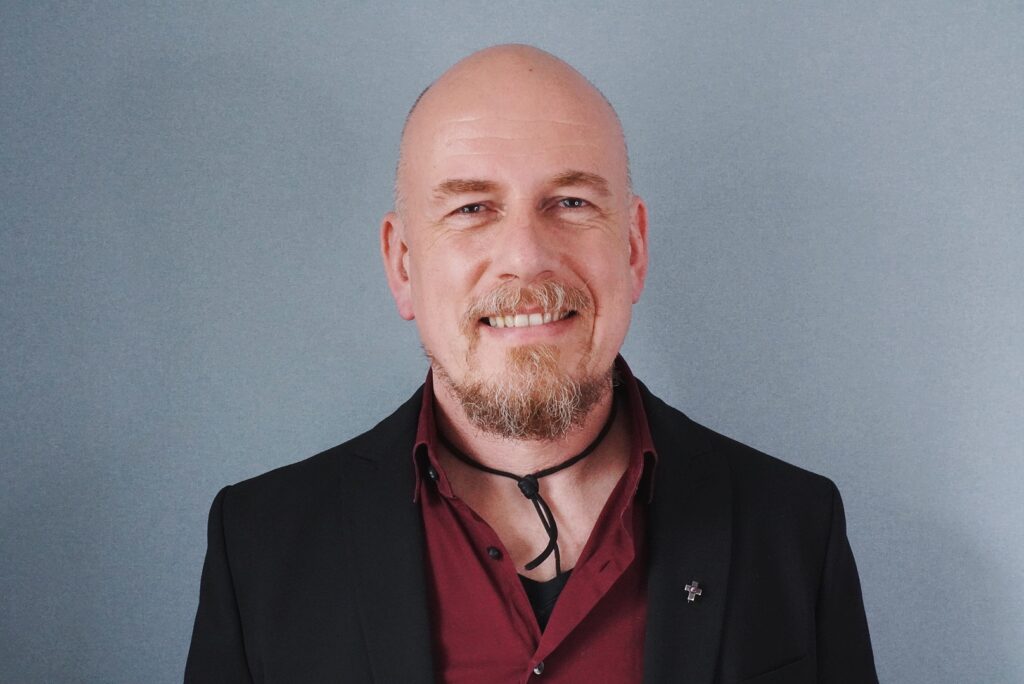 Es tut mir leid, ich weiß nicht, wer das ist. Hier ist ein Vorschlag für den Alt-Text: "Lächelnder Mann mit Glatze und Bart Porträt"