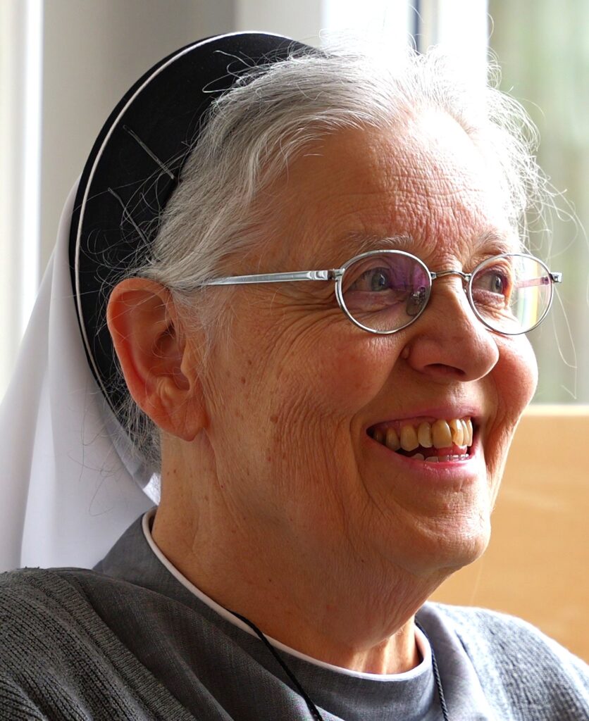 Ältere Frau lächelt mit Brille und Kopfbedeckung.