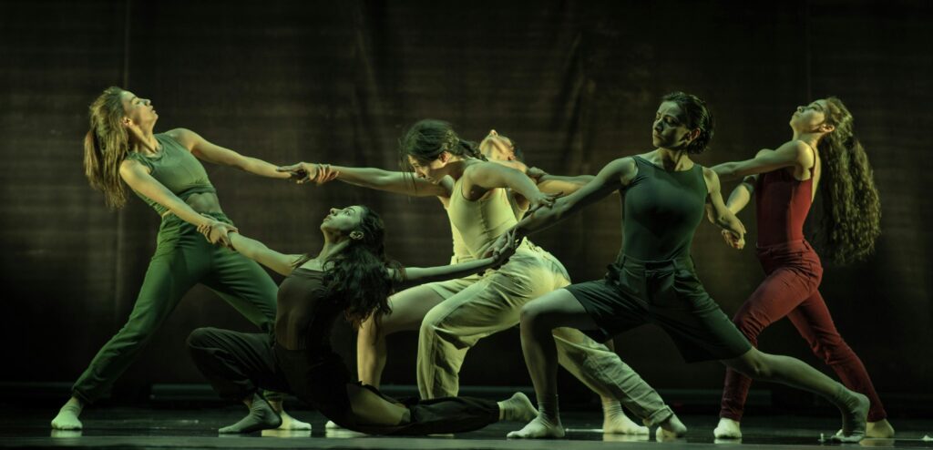 Sechs Tänzerinnen und Tänzer bilden in dramatischen Posen eine dynamische Gruppe auf der Bühne, halten sich an den Händen und strecken sich aus, tragen taillierte, erdfarbene Kostüme und werden von grünem Bühnenlicht begleitet.
