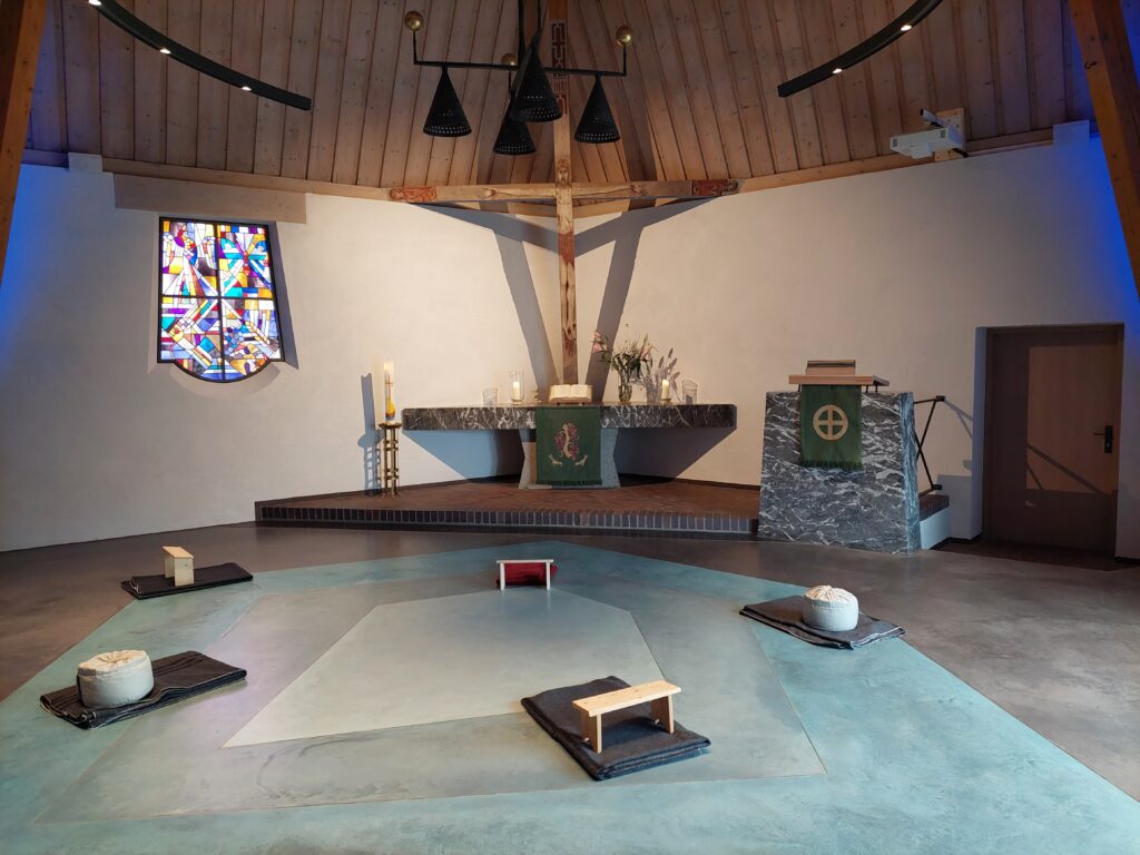 Ein modernes Kircheninnere mit einer Holzdecke, einem Altar, einem Kreuz, einem Buntglasfenster und Meditationskissen, die kreisförmig auf dem Boden angeordnet sind.