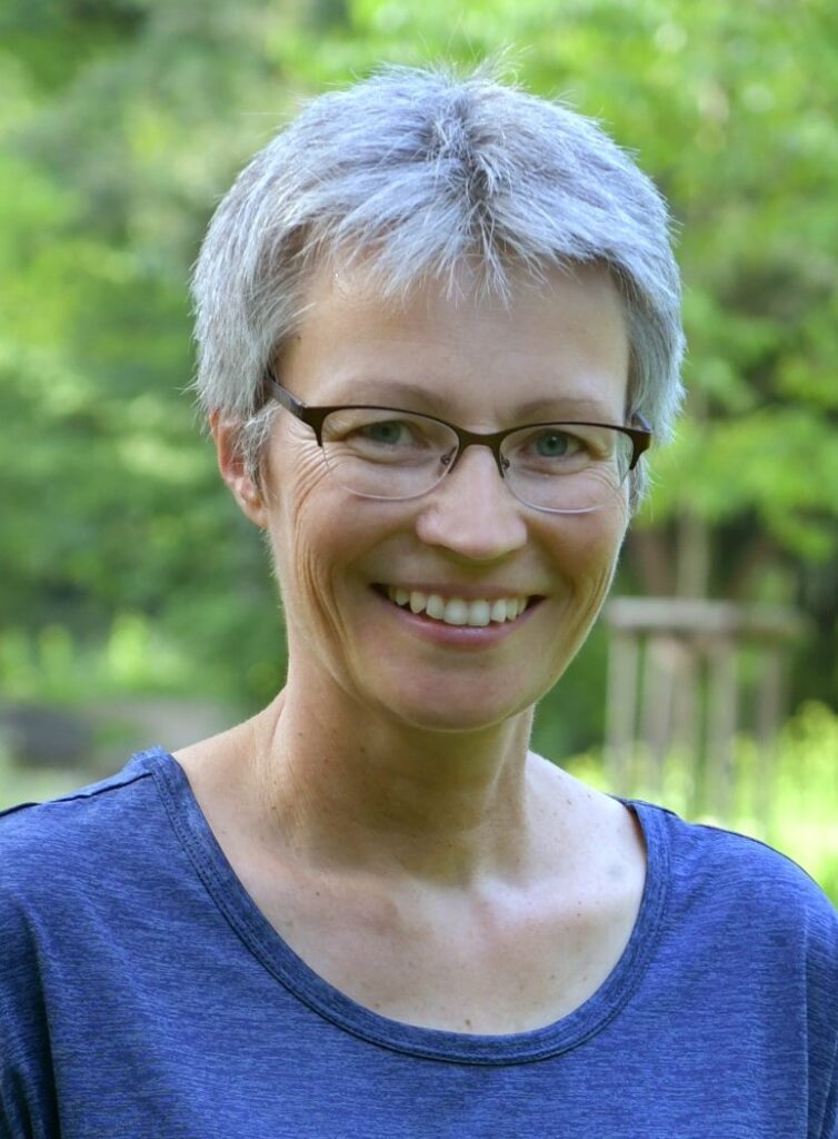 Lächelnde Person mit kurzen grauen Haaren und Brille, die ein blaues Oberteil trägt, steht im Freien mit grünen Bäumen im Hintergrund.