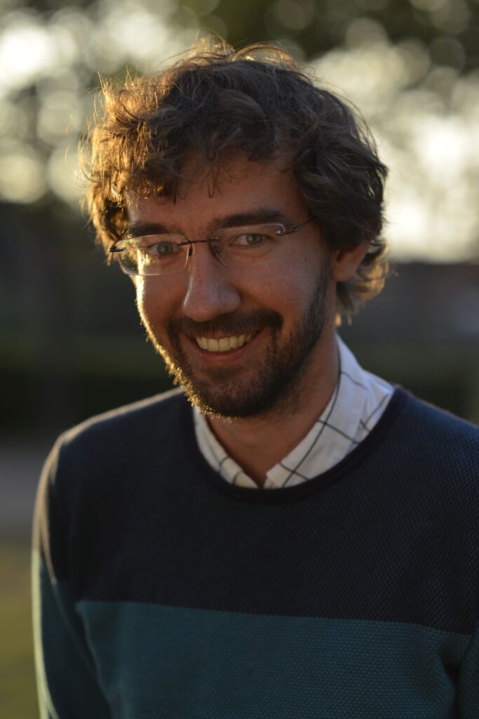 Ein Mann mit lockigen braunen Haaren, Brille und Bart lächelt draußen im warmen Sonnenlicht, er trägt einen Pullover über einem karierten Hemd.