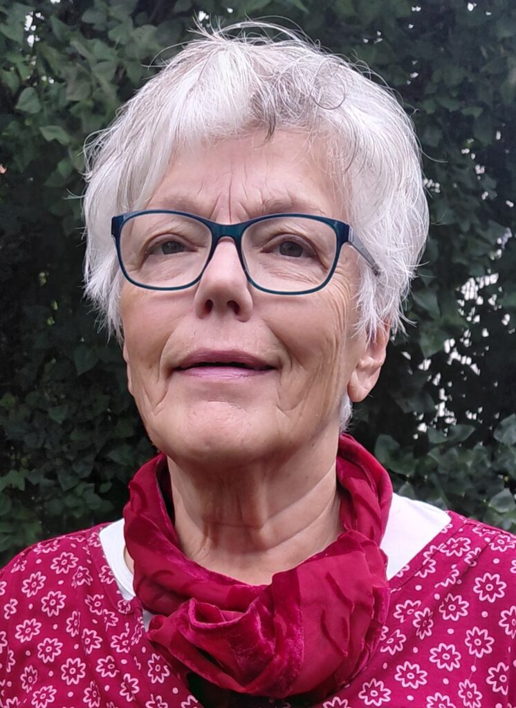 Ältere Frau mit kurzen weißen Haaren, Brille und rotem Schal, die im Freien vor grünem Laubgebüsch steht und sanft lächelt.