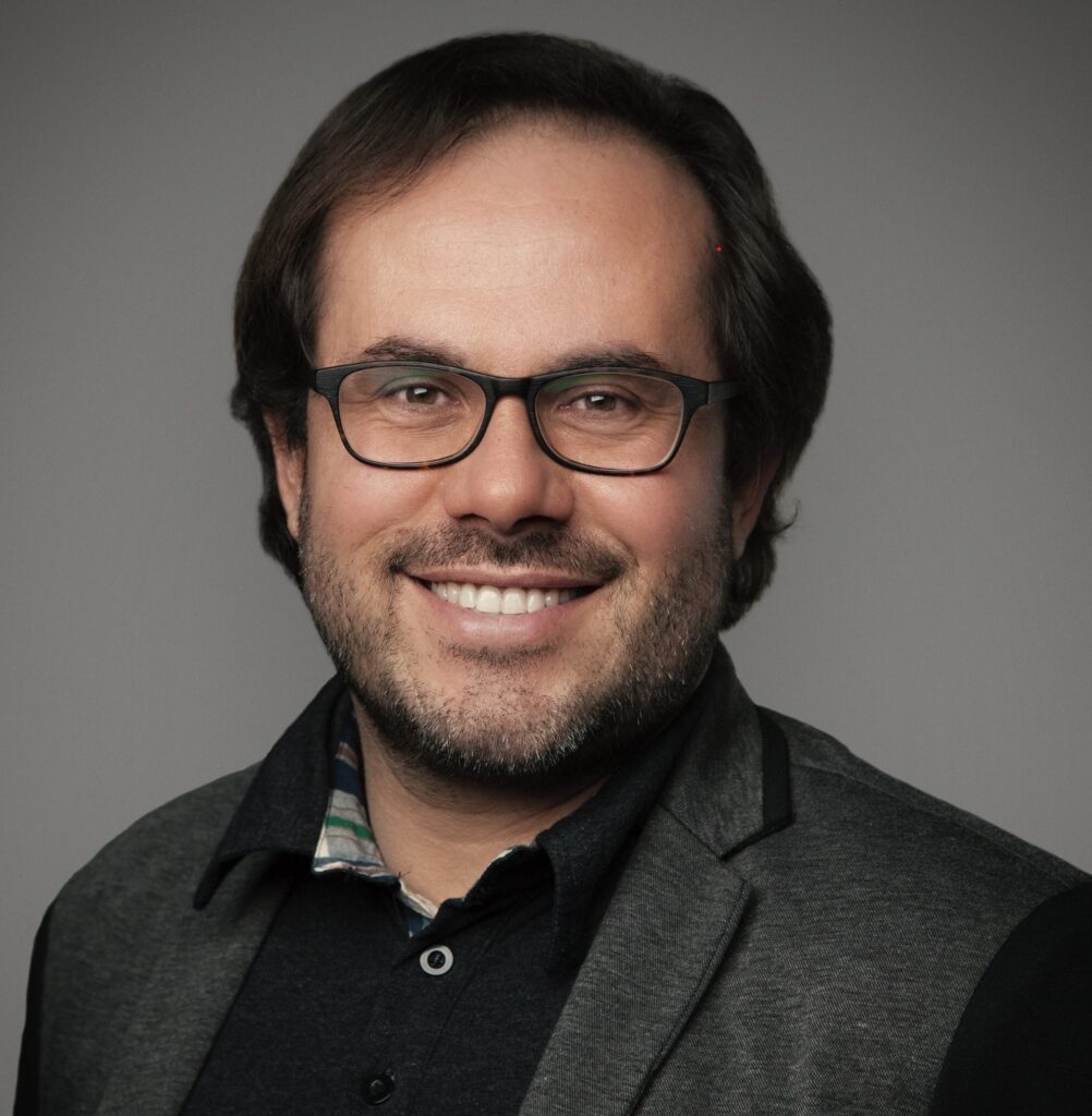 Lächelnde Person mit kurzen dunklen Haaren, die eine Brille, eine dunkle Jacke und ein schwarzes Hemd trägt, posiert vor einem schlichten hellen Hintergrund.