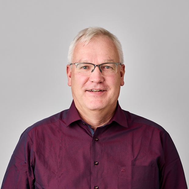 Ein lächelnder älterer Mann mit kurzen grauen Haaren und Brille, der ein burgunderrotes Button-up-Hemd trägt, steht vor einem schlichten hellgrauen Hintergrund.